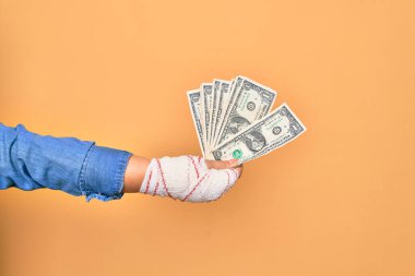 Kafkasyalı genç bir kadının yaralı eli, sargıyla kaplı, izole edilmiş sarı arka planda bir deste banknot tutuyor.