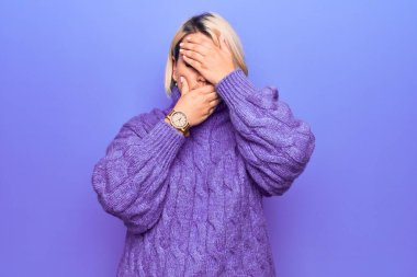 Güzel sarışın ve cüsseli bir kadın mor arka planda balıkçı yaka kazak giyiyor gözleri ve ağzı kapalı, şaşırmış ve şok olmuş. Duygularını gizliyor.