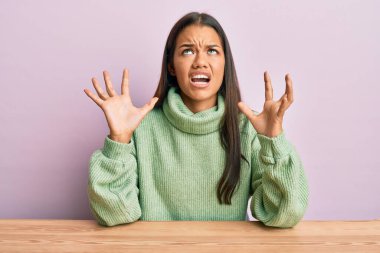 Güzel İspanyol kadın, gündelik giysiler içinde masada oturmuş deli gibi bağırıyor ve agresif bir ifadeyle bağırıyor ve kollarını kaldırıyor. hüsran kavramı. 