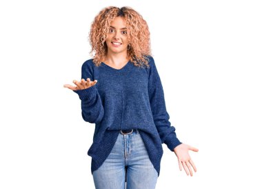 Kış süveteri giyen, kıvırcık saçlı genç bir kadın. Neşeli gülümsüyor, kollarını açıyor. Hoş geldin, olumlu ve kendinden emin selamlar. 