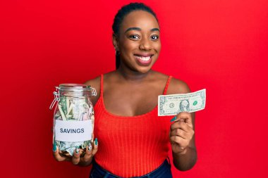 Genç Afro-Amerikalı bir kadın elinde kavanoz, elinde birikimleri ve yüzünde mutlu ve havalı bir gülümsemeyle gülümseyen bir banknot tutuyor. diş gösterme. 