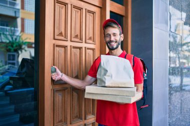 Beyaz teslimatçı, kırmızı üniforma giyiyor ve pizza kutusunu tutarken mutlu bir şekilde sırt çantası taşıyor.