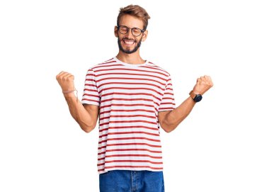 Sakallı, sıradan kıyafetler ve gözlük takan yakışıklı sarışın adam. Çok mutlu ve heyecanlı. Kolları havada, gülümseyen ve başarı için çığlık atan kazanan bir jest yapıyor. kutlama konsepti. 