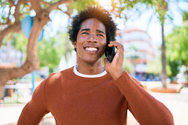 Genç, yakışıklı, Afro-Amerikan bir adam günlük giysiler giyip mutlu gülümsüyor. Yüzünde tebessümle dikilip şehrin caddesinde akıllı telefondan konuşuyor..