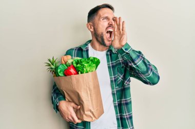 Elinde poşet sakallı yakışıklı bir adam alışveriş torbalarıyla bağırıyor ve ağızdan ağıza bağırıyor. iletişim konsepti. 