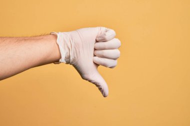 Sarı arka plan üzerinde tıbbi eldiveni olan beyaz bir gencin eli. Başparmak reddetme hareketi, onaylamama hareketi ve olumsuz işaret yapıyor.
