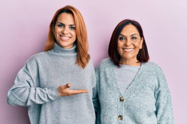 Latin anne ve kızı günlük kıyafetler giyip neşeli bir şekilde gülümsüyor ve avuç içiyle kameraya bakıyor.. 