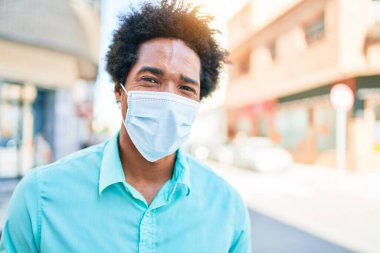 Genç, yakışıklı, Afro-Amerikan bir adam günlük kıyafetler ve tıbbi maske takıyor..