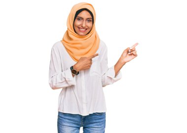 Genç esmer kadın geleneksel İslami başörtüsü takıyor gülümsüyor ve kameraya bakıyor iki eliyle ve parmaklarıyla yan tarafa bakıyor.. 