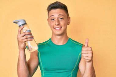Spor kıyafetleri giyen, elinde su şişesi tutan, mutlu ve pozitif gülümseyen, başparmağıyla mükemmel ve onay işareti yapan genç İspanyol çocuk. 