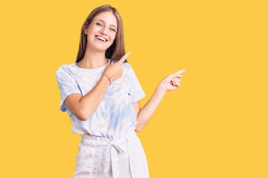 Tye tişörtü giyen genç ve güzel sarışın kadın gülümsüyor ve kameraya bakıyor iki eliyle ve parmaklarıyla kenarı işaret ediyor.. 