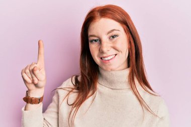 Balıkçı yaka kazak giyen genç kızıl saçlı kız bir fikir ya da soruyla gülümsüyor ve mutlu bir yüzle işaret ediyor. 