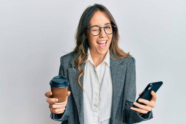 Genç esmer kadın akıllı telefon kullanıyor ve bir fincan kahve içiyor. Seksi bir ifadeyle kameraya bakıyor, neşeli ve mutlu bir yüzle.. 