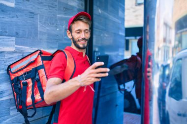 Beyaz teslimatçı kırmızı üniforma giyiyor ve sırt çantası teslim ederken gülümsüyor ve akıllı telefon kullanıyor.
