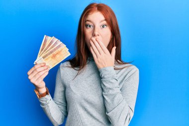 Young red head girl holding 500 norwegian krone banknotes covering mouth with hand, shocked and afraid for mistake. surprised expression 