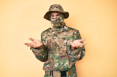 Kamuflaj üniforması giyen, kar maskesi takan, elleri ve kolları havada, kafası karışmış genç yakışıklı bir adam. şüphe kavramı. 