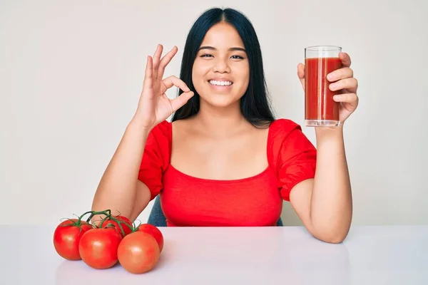 年轻美丽的亚洲女孩喝着一杯健康的番茄汁 用手指做着手势 微笑着表示友善的手势 是极好的象征 — 图库照片