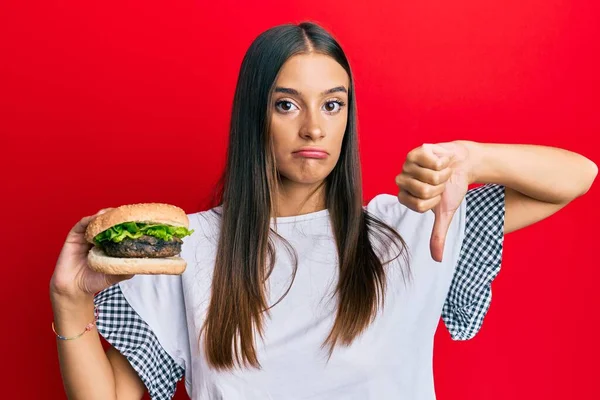 若いヒスパニック系の女性が怒っている顔でハンバーガーを食べます 親指で嫌いを示す負の兆候ダウン 拒絶概念 — ストック写真