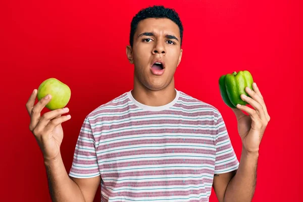 衝撃的な顔で緑のリンゴとコショウを保持若いアラブ人男性 懐疑的で皮肉を探して オープン口で驚いて — ストック写真