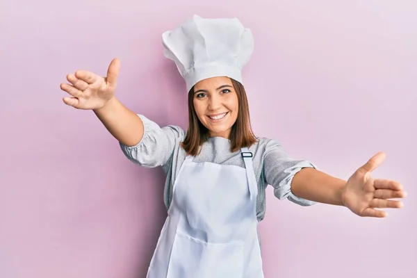年轻美丽的女人穿着职业厨师的制服和帽子 看着镜头 张开双臂拥抱 欢快的表达拥抱幸福 — 图库照片