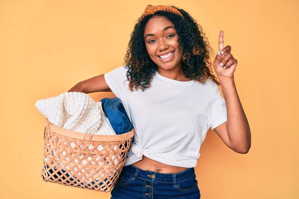 洗濯籠を持っている若いアフリカ系アメリカ人の女性は 幸せな顔 ナンバーワンで指を指しているアイデアや質問に驚いて — ストック写真