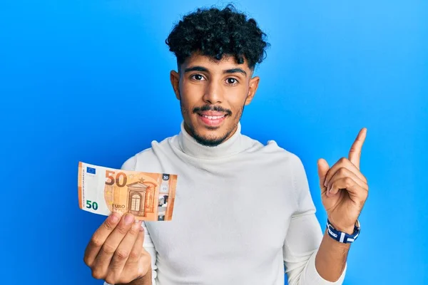 年轻的Arab帅哥拿着50欧元的钞票开心地微笑着 手指手画脚地指向旁边 — 图库照片