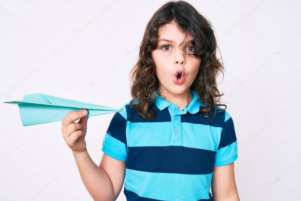 Cute hispanic child with long hair holding paper airplane scared and amazed with open mouth for surprise, disbelief face 