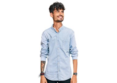 Gündelik kıyafetler giyen genç İspanyol bir adam yüzünde gülümsemeyle yan yana bakıyor, doğal bir ifade. Kendinden emin gülen. 