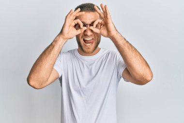 Günlük kıyafetler ve gözlük takan genç beyaz bir adam dürbün gibi jest yapıyor. Dürbün gibi dilini dışarı çıkarıyor, gözler parmaklarından bakıyor. çılgın ifade. 