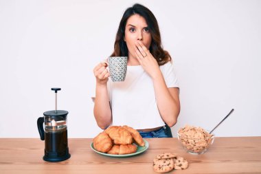 Genç, güzel esmer kadın masada oturmuş kahvaltısını yapıyor. Ağzını eliyle kapatıyor. Şok olmuş ve hata yapmaktan korkuyor. şaşırmış ifade 