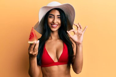 Güzel İspanyol kadın bikini giyiyor, karpuz yiyor parmaklarıyla işaret yapıyor, gülümseyerek mükemmel bir sembol oluşturuyor. 