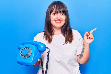 Elinde klasik bir telefon tutan genç ve cüsseli bir kadın gülümseyerek elini ve parmağını yana uzatarak mutlu mesut işaret ediyor. 