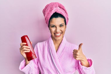 Duşlu bornoz giyen genç İspanyol kadın elinde şampuan, gülümsüyor mutlu ve pozitif, başparmağıyla mükemmel ve onay işareti yapıyor. 