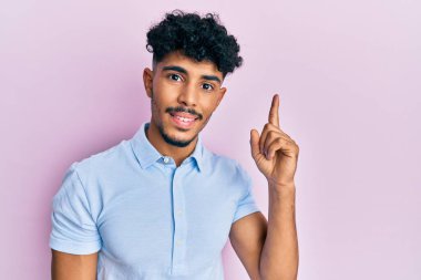 Gündelik giysiler giymiş genç Arap yakışıklı bir adam bir fikir veya soruyla gülümsüyor ve mutlu bir yüzle işaret ediyor. 