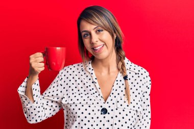 Genç ve güzel bir kadın kahve içerken pozitif ve mutlu görünüyor. Ayakta duruyor ve gülümsüyor. Dişlerini göstererek gülümsüyor. 