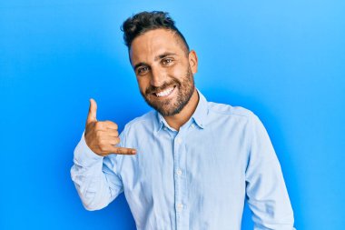 Sakallı, sıradan kıyafetler giyen, gülümseyen, el ve parmaklarıyla telefon hareketi yapan, telefonda konuşan yakışıklı bir adam. iletişim kavramları. 