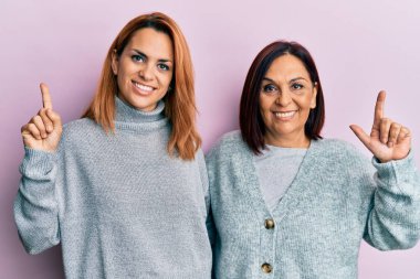 Latin anne ve kız, sıradan kıyafetler giyip, yüzünde kocaman bir gülümsemeyle, kameraya el ile işaret ediyorlar.. 