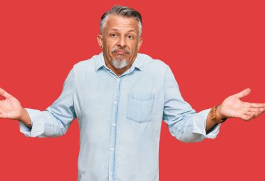 Orta yaşlı, gri saçlı, sıradan kıyafetler giyen, kafası karışmış, elleri ve kolları havada bir adam. şüphe kavramı. 