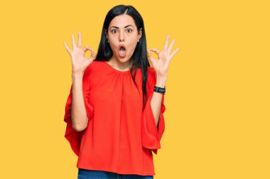 Gündelik kıyafetler giyen güzel genç bir kadın şaşırmış ve şok olmuş görünüyor. Tamam sembolünü parmaklarıyla yapıyor. çılgın ifade 