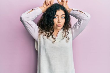 Genç esmer, kıvırcık saçlı, günlük kıyafetler giyen, boynuzlu parmaklarıyla komik jestler yapan bir kadın. 