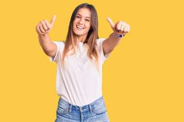 Güzel, beyaz tişört giyen beyaz bir kadın. Elle olumlu jest yapmayı onaylıyor. Başparmağıyla gülümsüyor ve başarı için seviniyor. kazanan jest. 