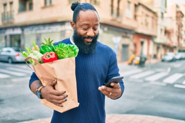 Sakallı Afrikalı Amerikalı adam elinde süpermarketten alınmış kağıt torbalarla akıllı telefon kullanıyor.