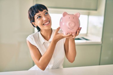 Genç esmer kadın mutlu bir şekilde gülümsüyor gururlu kumbarasını birikimleriyle gösteriyor