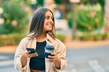 Genç İspanyol kız mutlu bir şekilde gülümsüyor. Akıllı telefon kullanıyor ve parkta kahve içiyor..