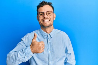 Gündelik giysiler ve gözlük takan İspanyol genç adam elleriyle mutlu bir jest yapıyor. Kameraya bakıp başarıyı gösteren ifade. 