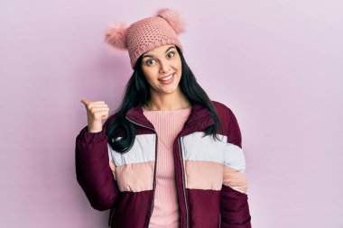 Genç İspanyol kadın yün kazak ve kep giyiyor, gülümsüyor, gülümsüyor ve başparmağıyla yan tarafa bakıyor.. 