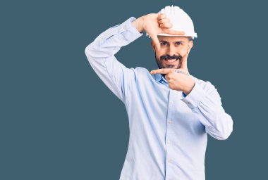 Mimar şapkası takan yakışıklı genç adam gülümsüyor. Elleriyle ve parmaklarıyla gülen yüzüyle çerçeve yapıyor. yaratıcılık ve fotoğrafçılık kavramı. 