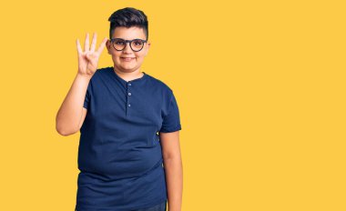 Küçük çocuk günlük kıyafetler ve gözlük takıyor. Dört numarayı gösteriyor ve işaret ediyor. Kendinden emin ve mutlu bir şekilde gülümsüyor.. 