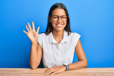 Güzel İspanyol kadın, gündelik giysiler içinde masada oturuyor ve beş numarayı göstererek gülümsüyor. Kendinden emin ve mutlu.. 