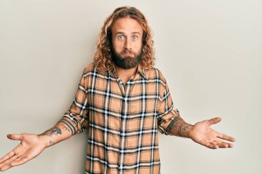 Sakallı, uzun saçlı, sıradan kıyafetler giyen, kafası karışmış ve kolları ve elleri havada yakışıklı bir adam. şüphe kavramı. 